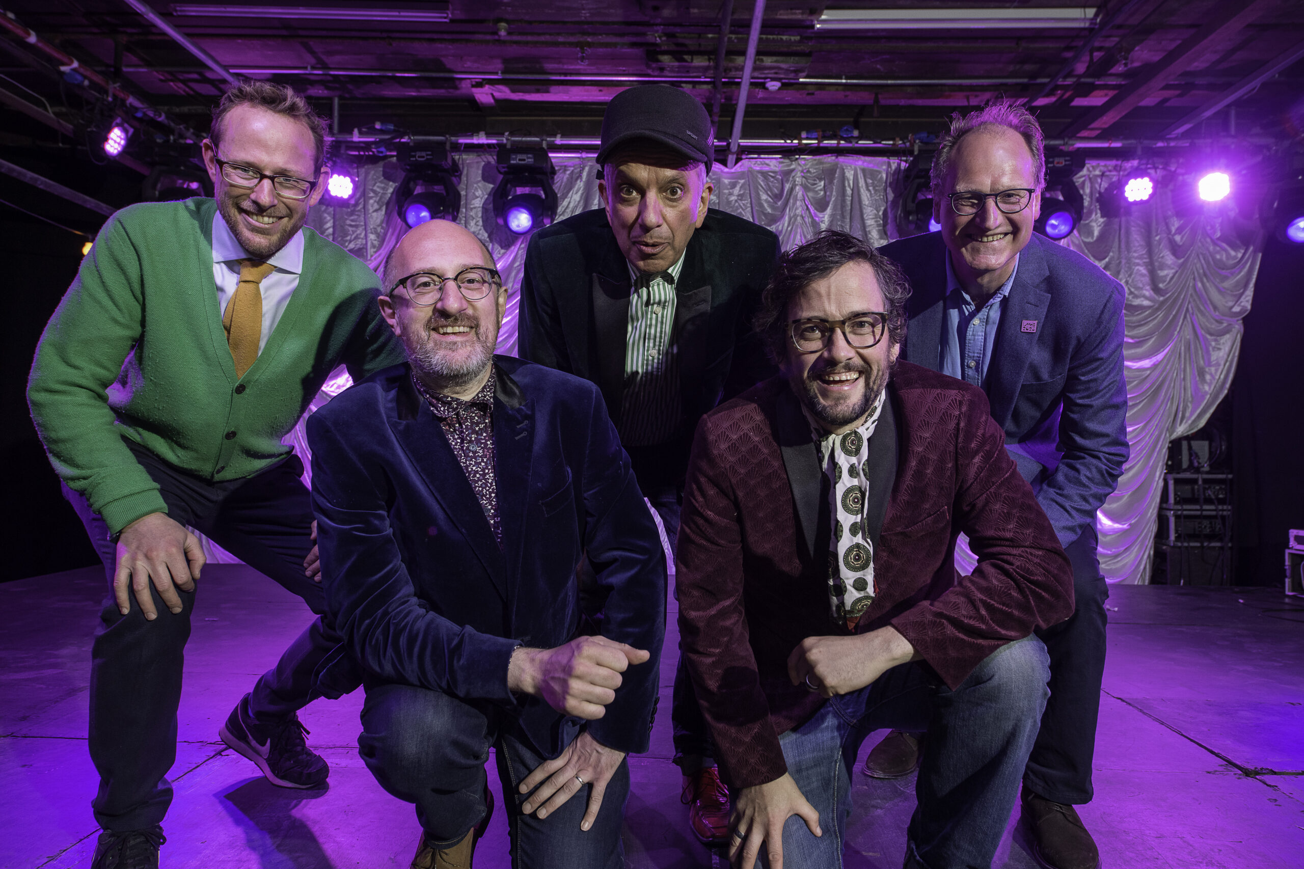 5 men crouched down and smiling at the camera inside a gig venue