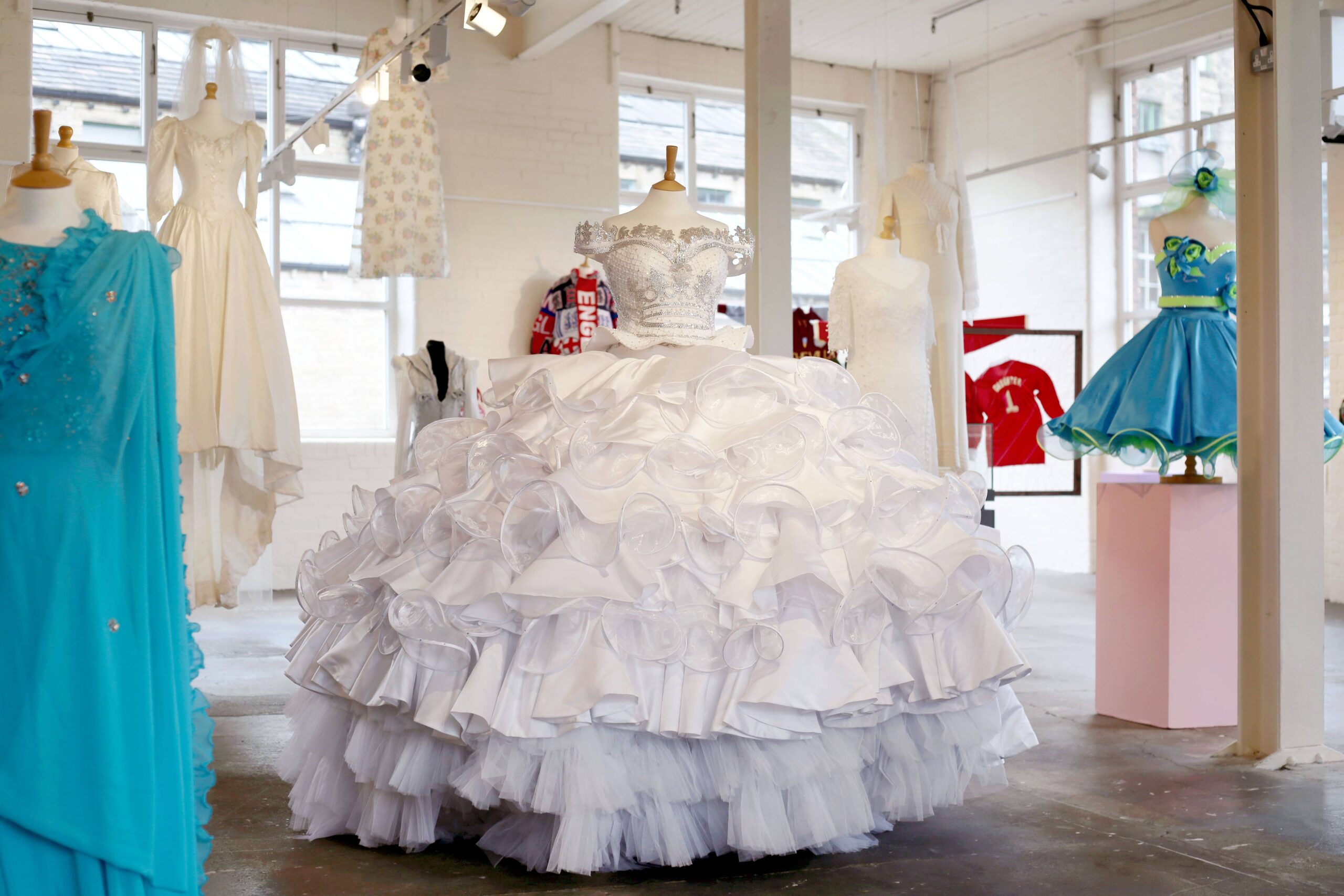 A white wedding dress with crystal and sequinned embellished bodice and a large many tiered skirt. There are other wedding outfits in the image.